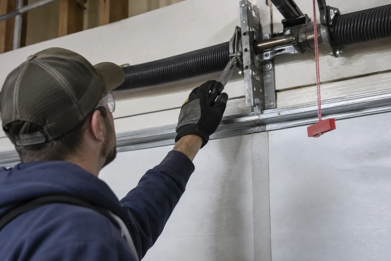 Technician in a cap reaching a gloved hand with a tool toward a coiled spring and bracket above a garage door.