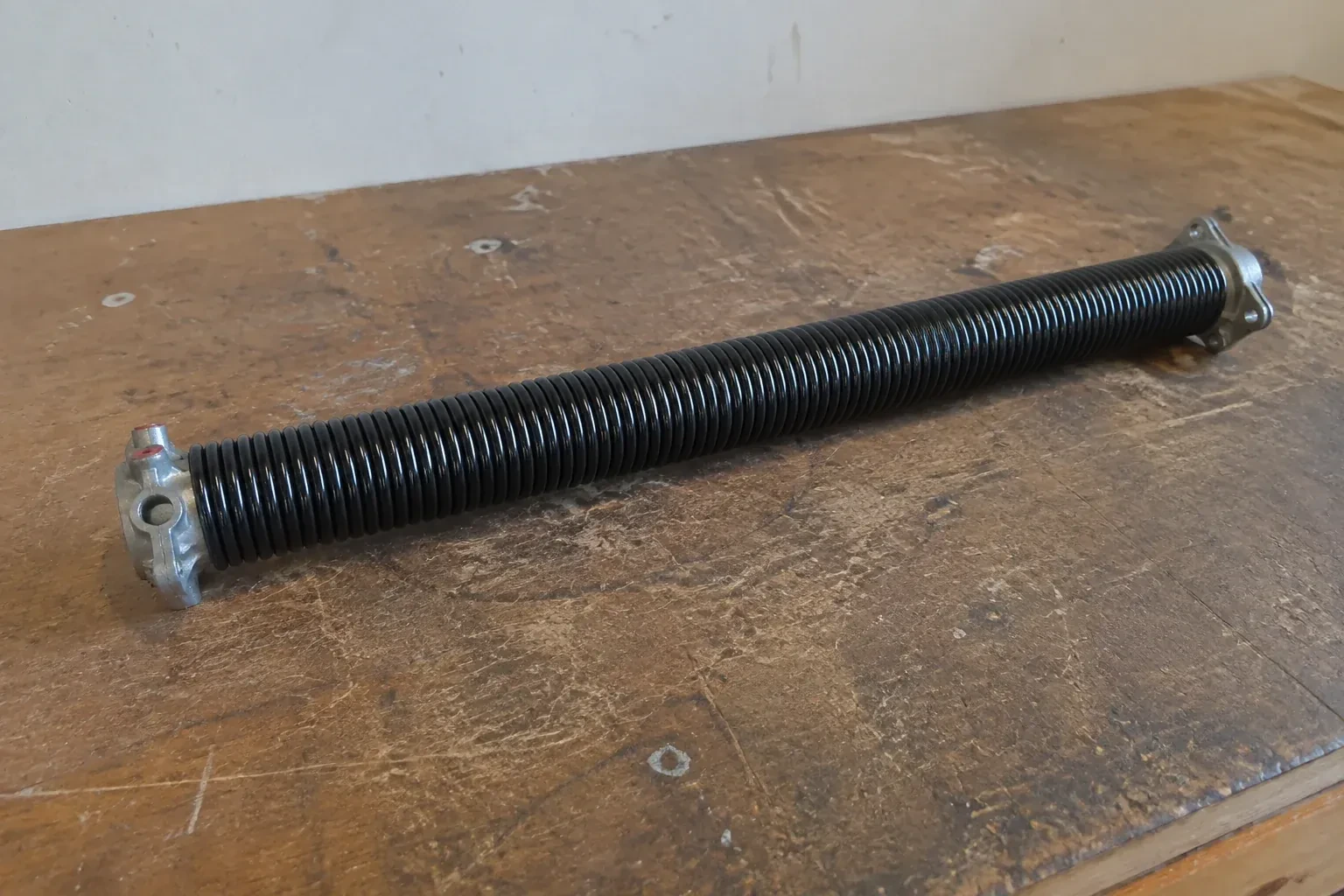 Detached black coil spring with metal end fittings on a workbench. — Torsion Spring Replacement, Richmond Hill